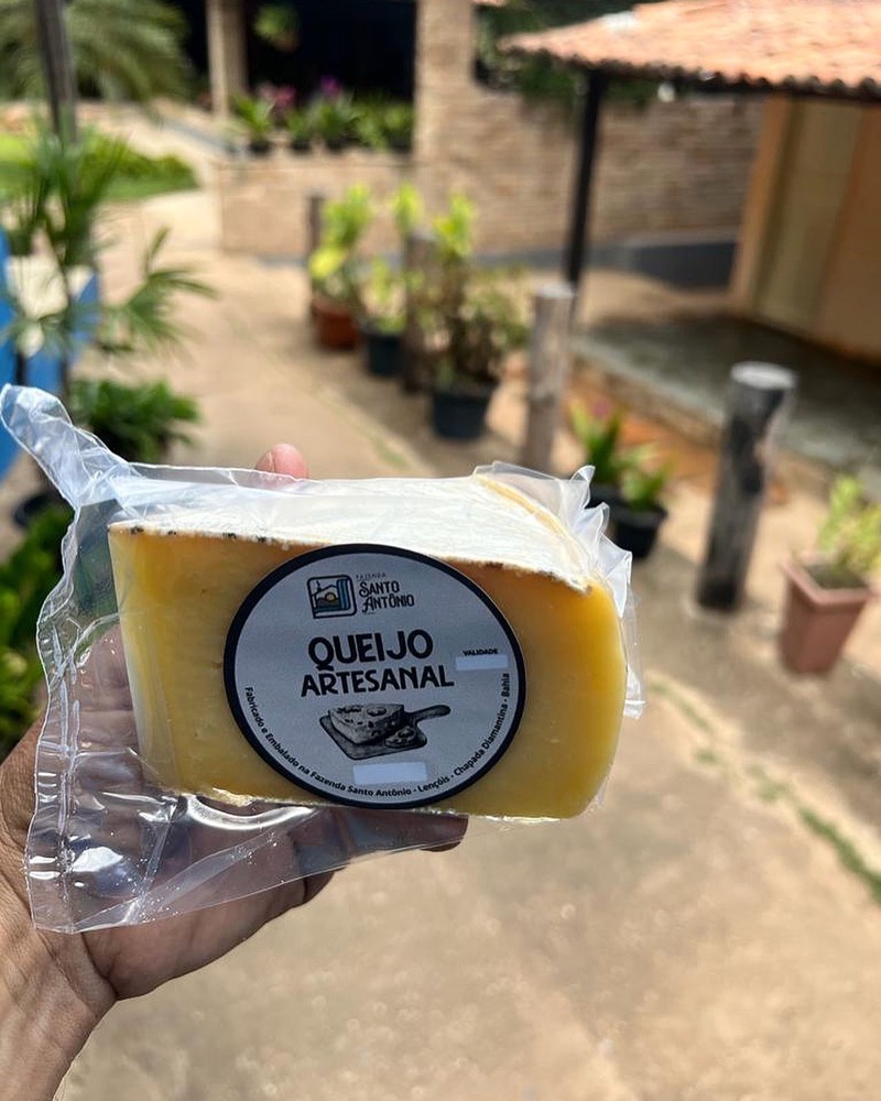 Mão segurando um pedaço de queijo artesanal embalado, com rótulo da Fazenda Santo Antônio e a área externa ao fundo